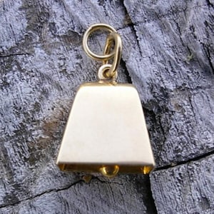 Pode incluir: Um pequeno pingente de sino em metal dourado com superfície lisa, formato trapezoidal e um anel circular na parte superior. O sino apresenta uma superfície simples e sem adornos, com um pequeno badalo visível no centro da abertura.