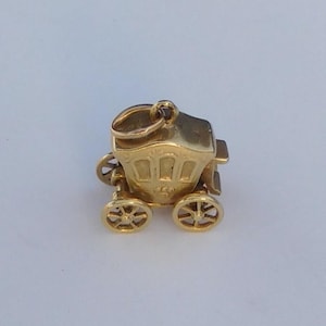 Op de afbeelding: Een kleine, gepolijste gouden bedel in de vorm van een koets. De bedel is voorzien van vier gedetailleerde spaakwielen, een rechthoekige koetsbak met boogvormige raamopeningen en een kleine ronde ring aan de bovenkant om hem op te hangen.