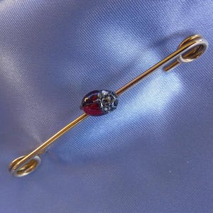 以下が含まれることがあります： 中央に楕円形のビーズが付いたゴールドトーンのバーピン。ビーズは深紅で、シルバーカラーのディテールが施されています。ピンは、明るいブルーのサテン生地の上に置かれています。