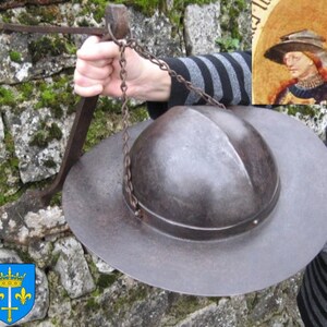 May include: A medieval helmet with a round, domed top and a wide brim. The helmet is made of metal and has a dark brown patina. It is hanging from a chain attached to a metal hook.