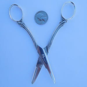 May include: A pair of silver-toned scissors with ornate handles, crossed and lying on a white surface. A US quarter is included for scale.