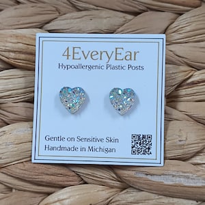 May include: A pair of heart-shaped stud earrings with a silver glitter finish on a white display card. The card features the text 4EveryEar, Hypoallergenic Plastic Posts, Gentle on Sensitive Skin, Handmade in Michigan, and a QR code.