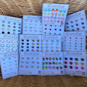 May include: A collection of 4EveryEar earring sets on white display cards. Each card features multiple pairs of hypoallergenic plastic stud earrings in various shapes, including hearts, stars, fruits, and geometric designs in many colors.