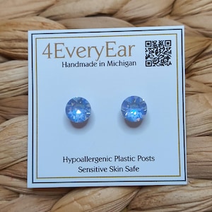 Puede incluir: Un par de pendientes de botón de cristal azul claro sobre una tarjeta de presentación blanca. La tarjeta incluye el texto 4EveryEar, Handmade in Michigan, un código QR y el texto Hypoallergenic Plastic Posts, Sensitive Skin Safe en negro.