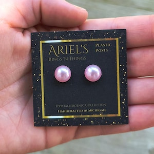 May include: A pair of pink pearl stud earrings on a black card with gold trim. The card reads "ARIEL'S RINGS 'N THINGS" and "PLASTIC POSTS". The card also states "HYPOALLERGENIC COLLECTION" and "HANDCRAFTED IN MICHIGAN."