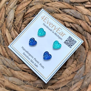 May include: Four heart-shaped stud earrings in shades of blue and teal, displayed on a white card. The card reads "4EveryEar Handmade in Michigan" and "Hypoallergenic Plastic Posts Sensitive Skin Safe". The earrings are on a woven background.
