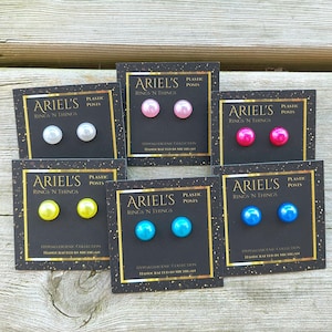 May include: A collection of colorful stud earrings on black cards. Each card features two round, plastic earrings in various colors: white, pink, hot pink, yellow, turquoise, and blue. The cards have gold borders and text that reads "ARIEL'S RINGS 'N THINGS".