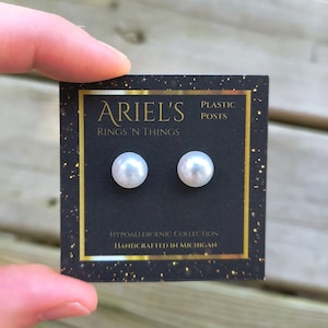 May include: A pair of white pearl stud earrings on a black card with gold trim. The card reads "ARIEL'S RINGS 'N THINGS" and "PLASTIC POSTS". The card also states "HYPOALLERGENIC COLLECTION HANDCRAFTED IN MICHIGAN".