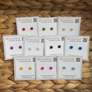 May include: A collection of nine pairs of stud earrings on white display cards labeled 4EveryEar Handmade in Michigan. Each pair features small, round, solid-colored spheres in red, green, yellow, purple, white, black, blue, pink, and light blue.