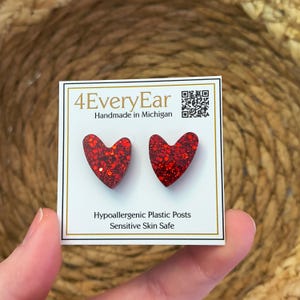 May include: A pair of red heart-shaped earrings with glitter, displayed on a white card with the text "4EveryEar Handmade in Michigan". The card also states "Hypoallergenic Plastic Posts Sensitive Skin Safe".