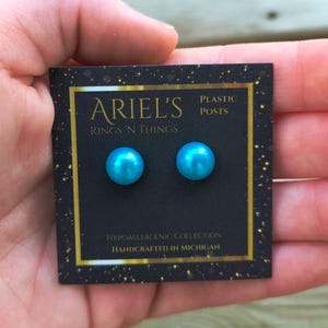May include: A pair of light blue pearl earrings on a black card with gold trim. The card reads "ARIEL'S RINGS 'N THINGS" and "PLASTIC POSTS". The earrings are part of the "HYPOALLERGENIC COLLECTION" and "HANDCRAFTED IN MICHIGAN".
