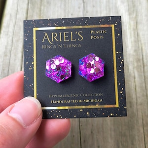 May include: A pair of hexagonal purple glitter earrings on a black card. The card reads "ARIEL'S RINGS 'N THINGS" and "HYPOALLERGENIC COLLECTION HANDCRAFTED IN MICHIGAN". The earrings have plastic posts.