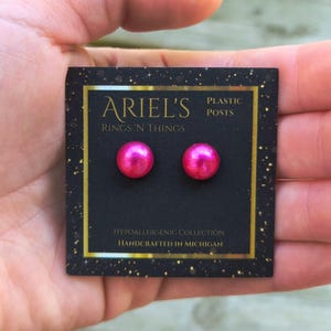 May include: A pair of bright pink, round stud earrings on a black card. The card has gold lettering that reads "ARIEL'S RINGS 'N THINGS" and "PLASTIC POSTS". The card also says "HYPOALLERGENIC COLLECTION" and "HANDCRAFTED IN MICHIGAN".