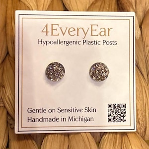 May include: A pair of round, silver-toned stud earrings with a textured, glittery surface. The earrings are mounted on a white display card labeled 4EveryEar, Hypoallergenic Plastic Posts, Gentle on Sensitive Skin, and Handmade in Michigan.