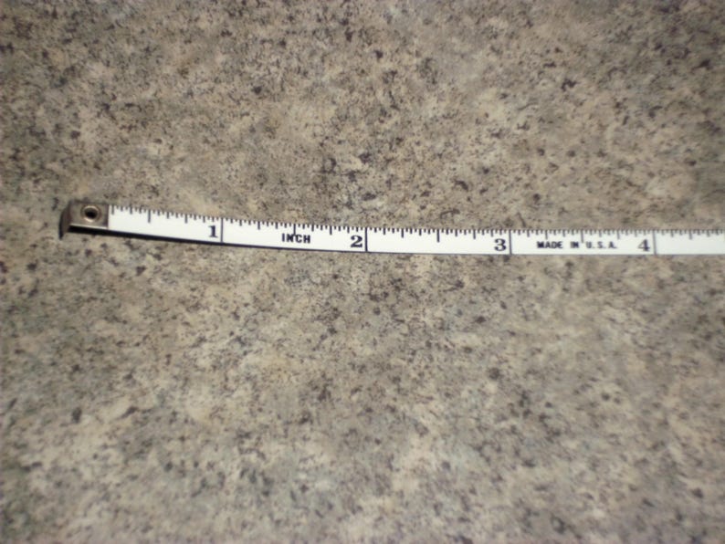 Pu&ograve; includere: Un metro a nastro bianco con segni neri, che mostra pollici. Il nastro misura fino a 10 cm. Il nastro &egrave; su una superficie strutturata dai toni neutri.