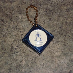 Pu&ograve; includere: Un portachiavi vintage blu e bianco con una custodia a forma di diamante e un inserto rotondo. L'inserto mostra un'illustrazione blu di una figura e il testo "NY YORK HANDER". Il portachiavi ha una catena a sfera color oro.