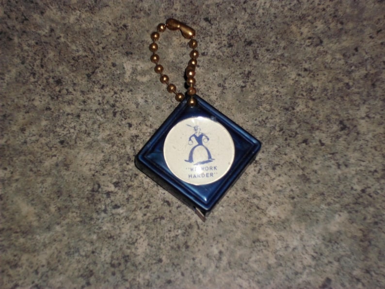 Pu&ograve; includere: Un portachiavi vintage blu e bianco con una custodia a forma di diamante e un inserto rotondo. L'inserto mostra un'illustrazione blu di una figura e il testo "NY YORK HANDER". Il portachiavi ha una catena a sfera color oro.