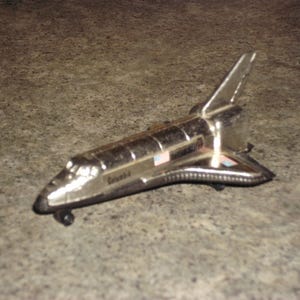 May include: A silver-colored toy space shuttle with the word "Columbia" printed on the side. The toy has a detailed design, including wings, a tail fin, and small wheels. The shuttle features American flags.
