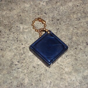 Pu&ograve; includere: Un piccolo oggetto blu navy a forma di diamante con una catena a sfera color oro. L'oggetto ha una superficie liscia e lucida e un bordo rialzato. La catena &egrave; attaccata alla parte superiore dell'oggetto.