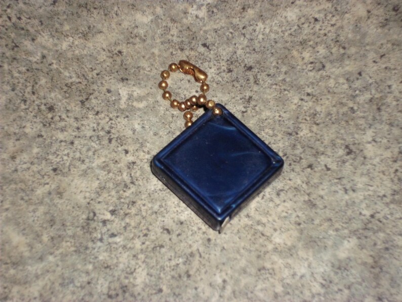 Pu&ograve; includere: Un piccolo oggetto blu navy a forma di diamante con una catena a sfera color oro. L'oggetto ha una superficie liscia e lucida e un bordo rialzato. La catena &egrave; attaccata alla parte superiore dell'oggetto.