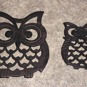 May include: Two cast iron owl-shaped trivets. The larger trivet has detailed feather and eye designs. The smaller trivet has a simpler design. Both are dark brown and sit on a gray surface. These trivets are decorative and functional.
