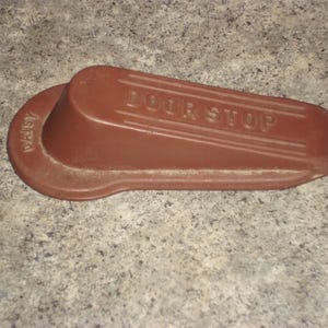 May include: A brown doorstop with the word "DOOR STOP" embossed on the top. The doorstop has a rounded end with the name "DAISY" printed on it. The doorstop is made of a solid material and is designed to prevent a door from opening too far.
