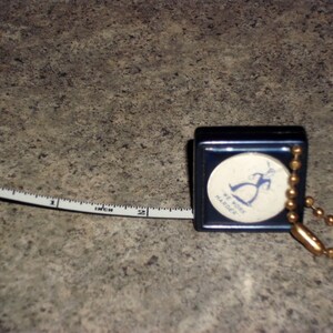 Pu&ograve; includere: Un metro a nastro blu navy con un metro a nastro bianco e nero esteso a 5 cm. Il metro a nastro ha una catena dorata e un emblema circolare con una grafica blu e la scritta "WE WORK HARDER."