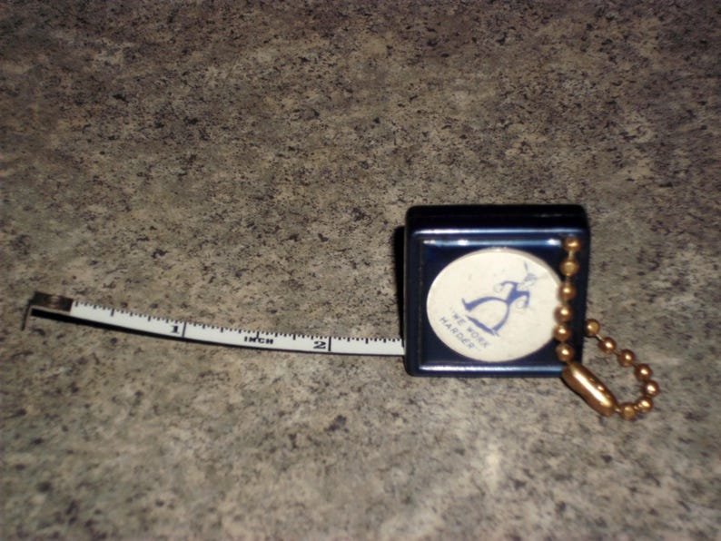 Pu&ograve; includere: Un metro a nastro blu navy con un metro a nastro bianco e nero esteso a 5 cm. Il metro a nastro ha una catena dorata e un emblema circolare con una grafica blu e la scritta "WE WORK HARDER."