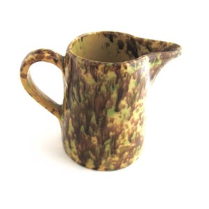 May include: A small, ceramic pitcher with a handle and spout. The pitcher has a speckled pattern in shades of brown, green, and yellow. The handle and spout are a solid yellow color. The pitcher is likely used for serving liquids.