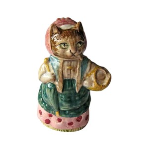 May include: A ceramic figurine of a cat wearing a pink bonnet and a green dress. The cat is holding a basket in one hand and a stick in the other. The figurine is painted with brown, green, pink, and yellow colors.