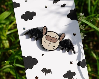 Puede incluir: Un pin de esmalte con forma de murciélago con alas negras y cara beige, sobre una tarjeta blanca con diseños de nubes y estrellas negras. La tarjeta también presenta el texto "noristudio". El pin se muestra sobre hierba verde.