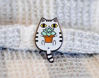 May include: A white cat-shaped enamel pin with black stripes and a black outline. The cat holds a small brown pot with a green succulent plant. The cat has yellow eyes and a striped tail. The pin is on a textured, light-coloured surface.