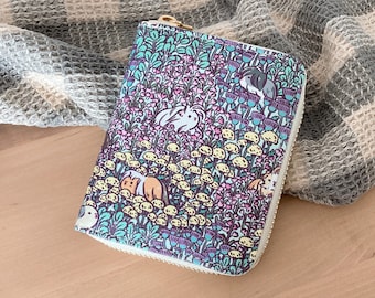 May include: A zippered wallet with a colorful pattern of rabbits, flowers, and mushrooms. The wallet has a white zipper and a gold-colored zipper pull. The background is a gray and white checkered fabric.