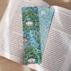 May include: Three decorative bookmarks with floral and animal designs. One bookmark features a pattern of light blue flowers and white cloud-like creatures. Another has a light blue floral pattern with a white animal. The third bookmark has a pattern of yellow flowers and a brown animal.