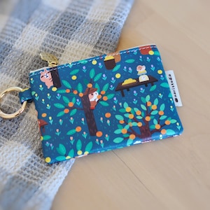 May include: A teal coin purse with a whimsical illustration of orange trees, animals, and a table. The purse has a gold zipper and a gold key ring. The brand name "@moristudio" is printed on a white tag.