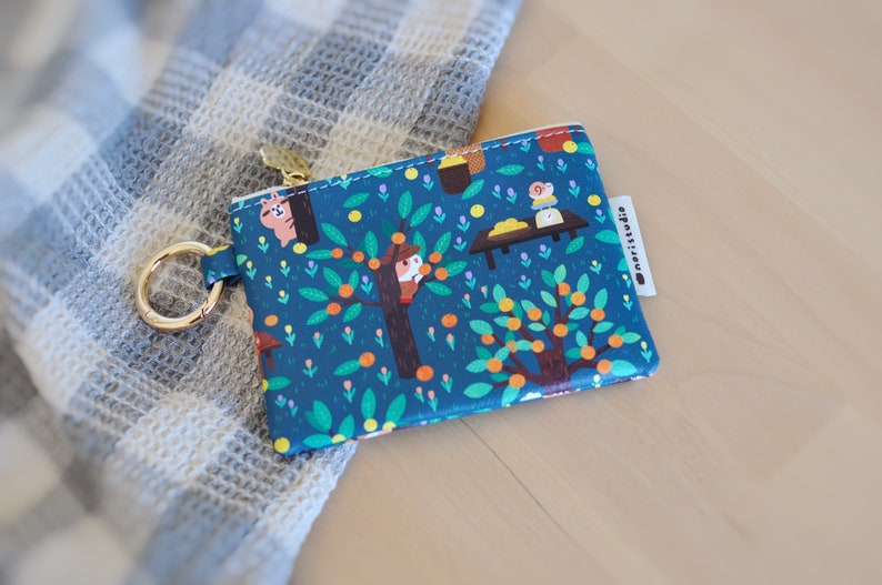 May include: A teal coin purse with a whimsical illustration of orange trees, animals, and a table. The purse has a gold zipper and a gold key ring. The brand name "@moristudio" is printed on a white tag.