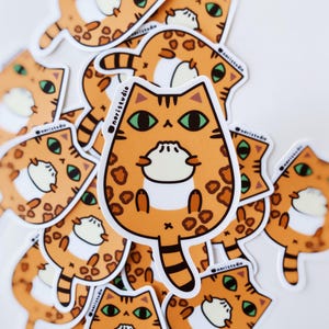Pode incluir: Autocolantes de gatos laranja com manchas pretas e olhos verdes espalhados numa superfície branca. Os autocolantes têm um design de desenho animado com uma barriga branca e o texto "@naristudio".