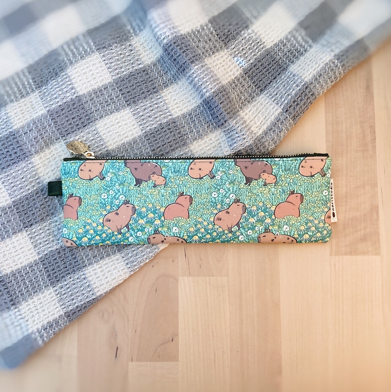 May include: A rectangular pencil case with a teal background featuring a repeating pattern of capybaras and small white and yellow flowers. The pencil case has a black zipper and a small green tab on one end.