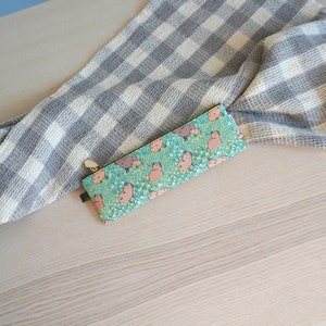 May include: A teal pencil case with a capybara pattern, featuring pink capybaras and yellow flowers. A gray and white checkered towel is draped in the background. The pencil case has a green tag and a gold zipper.