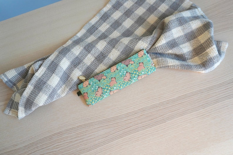 May include: A teal pencil case with a capybara pattern, featuring pink capybaras and yellow flowers. A gray and white checkered towel is draped in the background. The pencil case has a green tag and a gold zipper.