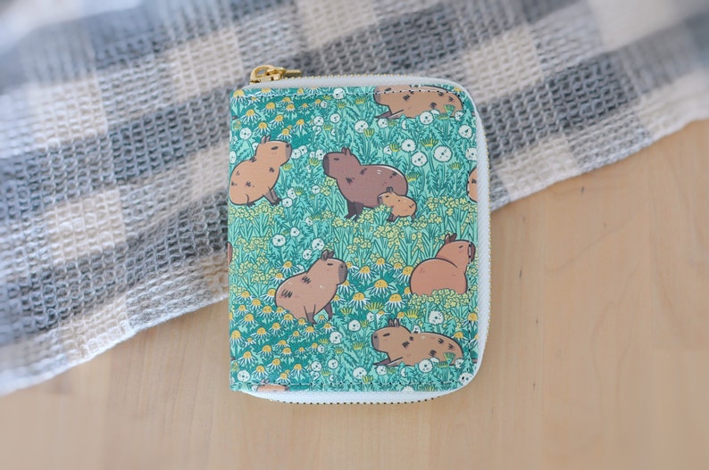 Puede incluir: Una cartera con cremallera con un patr&oacute;n repetido de carpinchos en un campo de margaritas. La cartera tiene un fondo verde azulado, una cremallera dorada y ribetes blancos. Los carpinchos son de varios tonos marrones.
