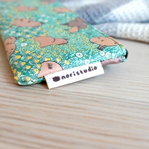 May include: A small, patterned pouch featuring a repeating design of brown animals on a green floral background. The pouch has a white tag that reads "noristudio" in black text. The background is a light wooden surface.
