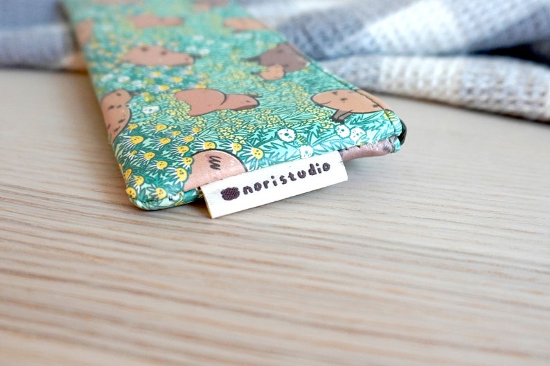 May include: A small, patterned pouch featuring a repeating design of brown animals on a green floral background. The pouch has a white tag that reads "noristudio" in black text. The background is a light wooden surface.
