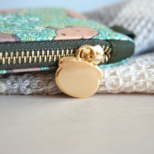 May include: Close-up of a small, zippered wallet with a gold-colored zipper and pull. The wallet features a green and brown pattern with a gold bear-shaped zipper pull. The background is a soft, textured fabric.