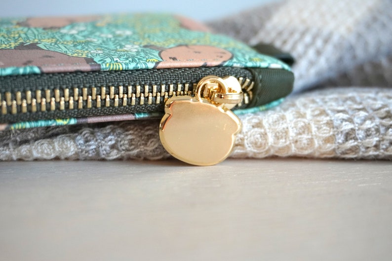May include: Close-up of a small, zippered wallet with a gold-colored zipper and pull. The wallet features a green and brown pattern with a gold bear-shaped zipper pull. The background is a soft, textured fabric.