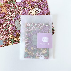 May include: A floral print fabric square with purple flowers and white rabbits is partially visible through a translucent white envelope. A matching floral print card with rabbit illustrations is in the background. A small purple sticker with the text "noristudio" is on the envelope.