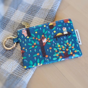 May include: A small, rectangular coin purse with a whimsical illustration of a tree with orange fruits, animals, and a table scene on a dark teal background. It has a gold-colored zipper and a matching key ring. The brand name "noristudio" is printed on a white tag.