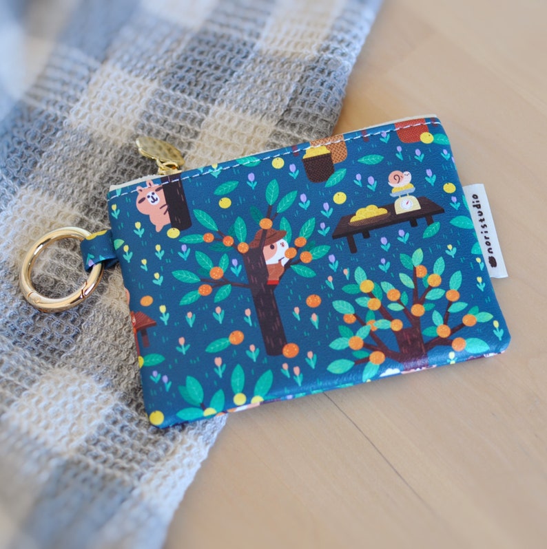 May include: A small, rectangular coin purse with a whimsical illustration of a tree with orange fruits, animals, and a table scene on a dark teal background. It has a gold-colored zipper and a matching key ring. The brand name "noristudio" is printed on a white tag.