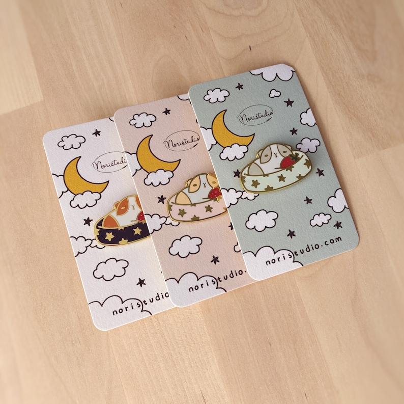 May include: Three enamel pins featuring a guinea pig in a bed with a strawberry, set on cards with a moon and star design. The pins have gold-colored borders and are on cards with the text "noristudio.com". The cards have a white, pink, and green background.