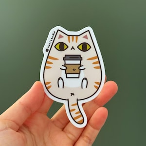 May include: A sticker featuring a cartoon cat holding a coffee cup. The cat is light beige with orange stripes and yellow eyes. The coffee cup is brown and white. The sticker has the text "noristudio" on the top left.